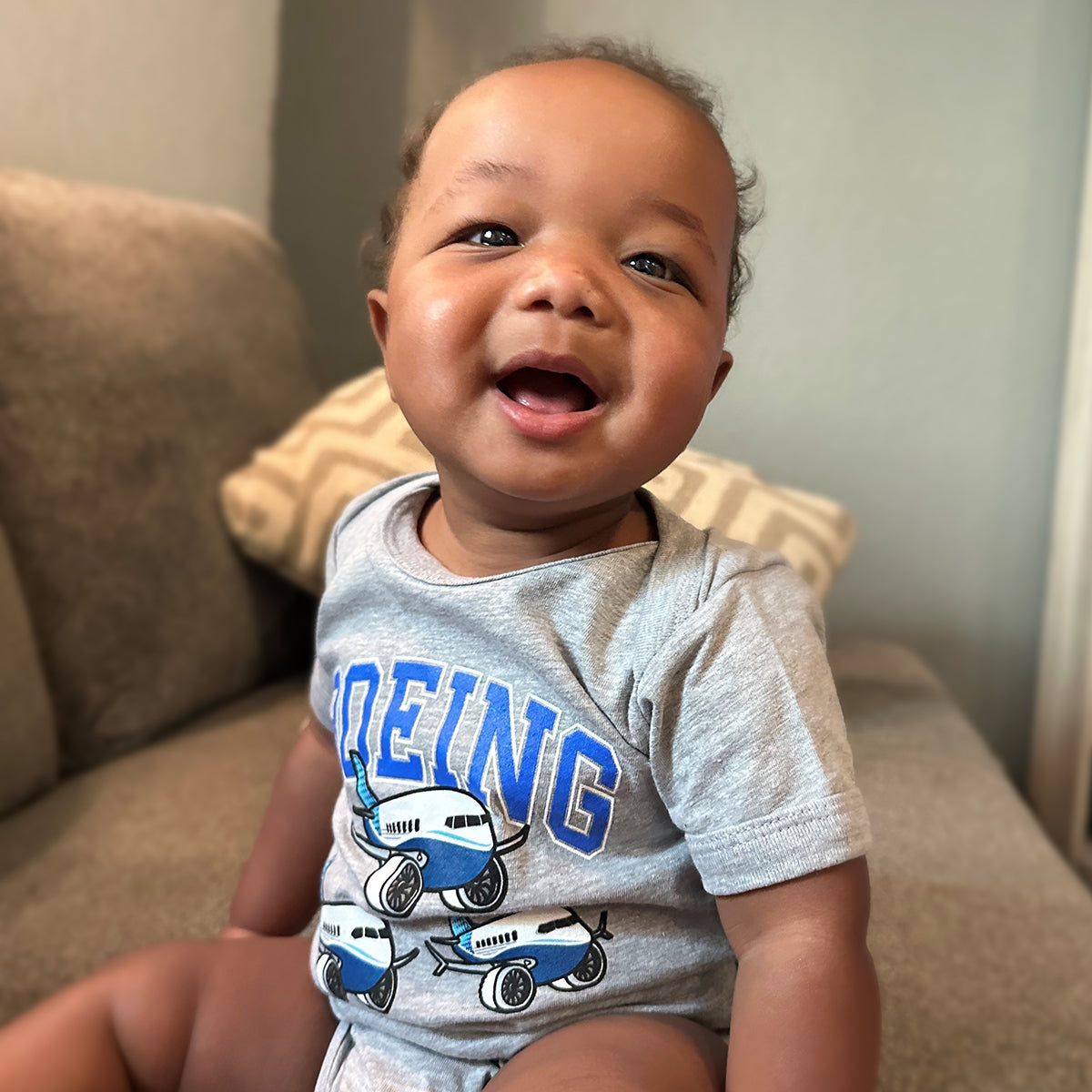 A smiling baby, on a couch, wears the Boeing Baby Varsity Pudgy Onesie crafted from sustainable cotton. The gray onesie has blue and white airplane graphics with BEING text. A beige pillow and soft-focus wall enhance the scene.