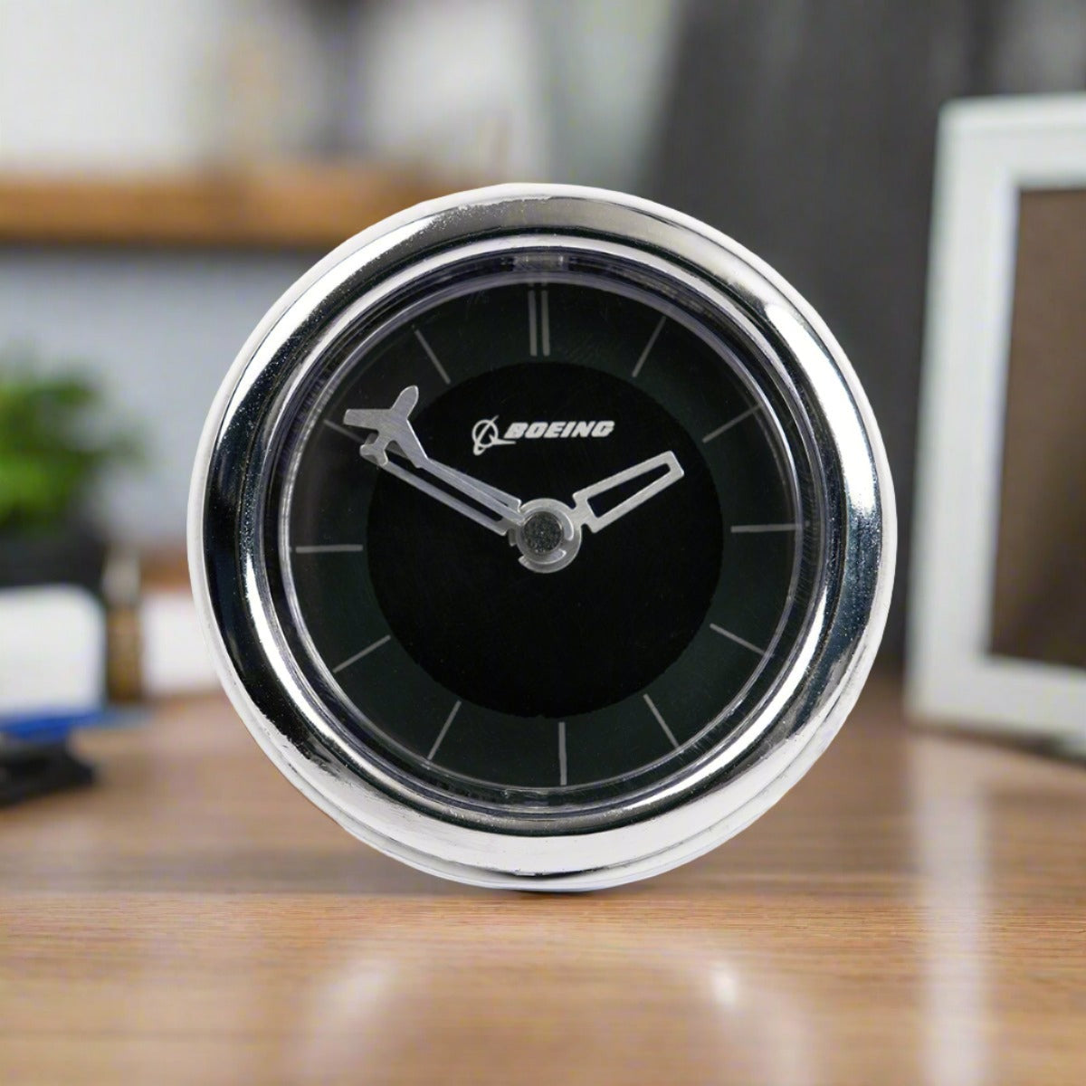 The Boeing Airplane Engine Desktop Clock by Boeing features a metallic frame, black face with a white Boeing logo, and airplane-shaped hour and minute hands. Perfect for aviation fans, its shown on a wooden surface with blurred office items in the background.