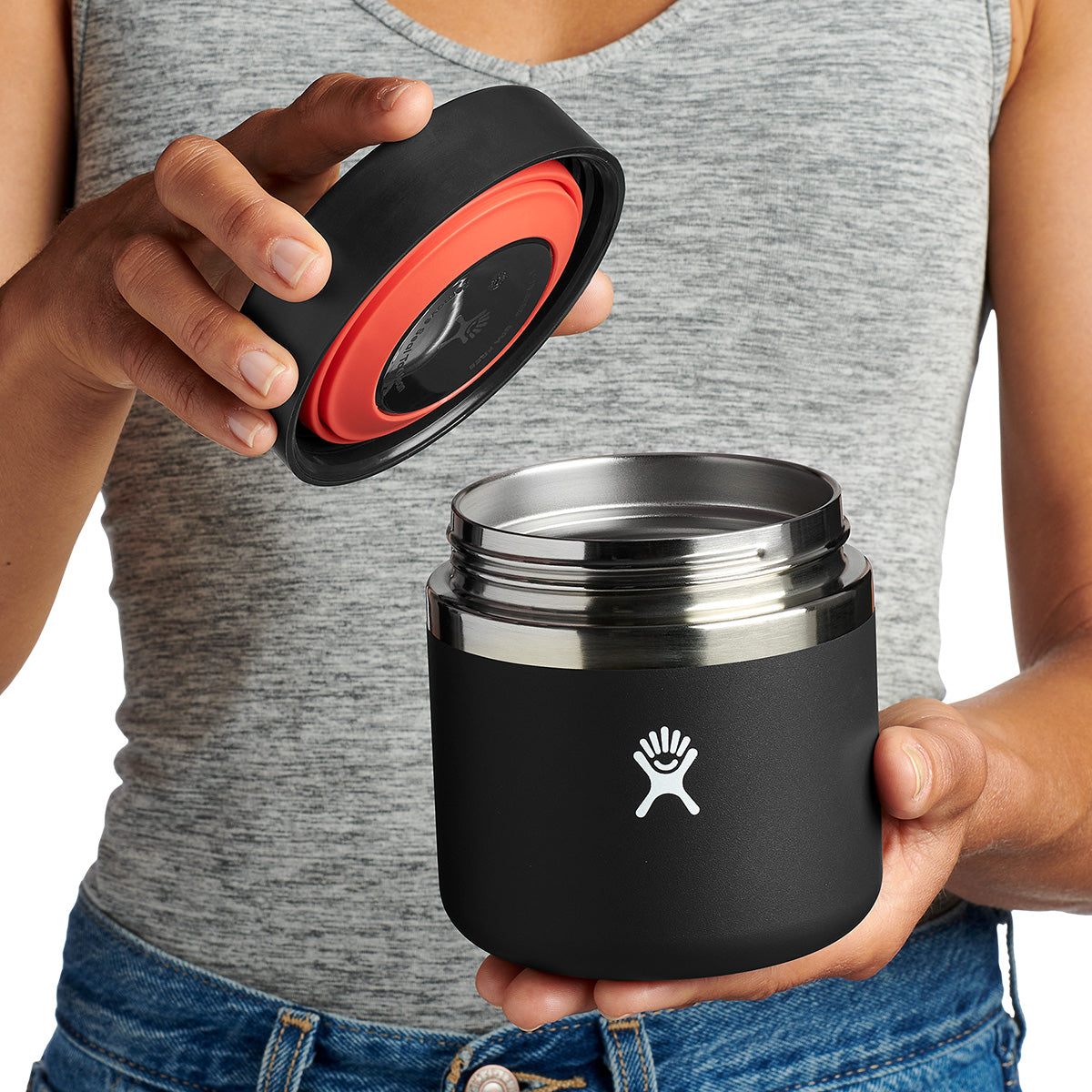 A person in a gray tank top holds a black Hydroflask Boeing 20 oz Food Jar with a silver rim, unscrewing the lid with both hands. The jar displays a white logo and features TempShield™ insulation by BOEING for lasting freshness.