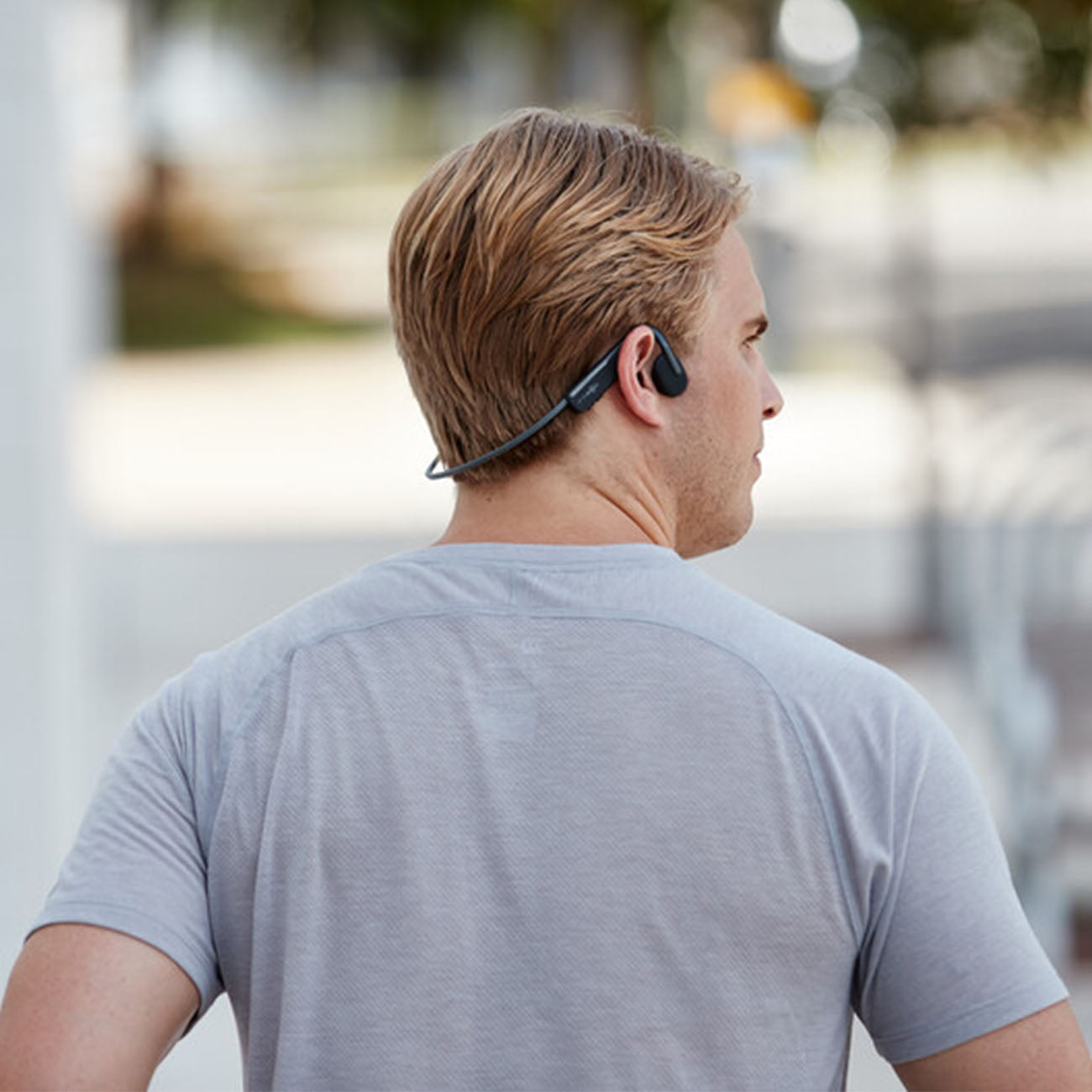 Outdoors against a blurred backdrop of trees and pavement, a blonde person wearing a gray T-shirt has Boeings Aftershokz Open Move Headphones with bone conduction technology around their neck, facing away from view.