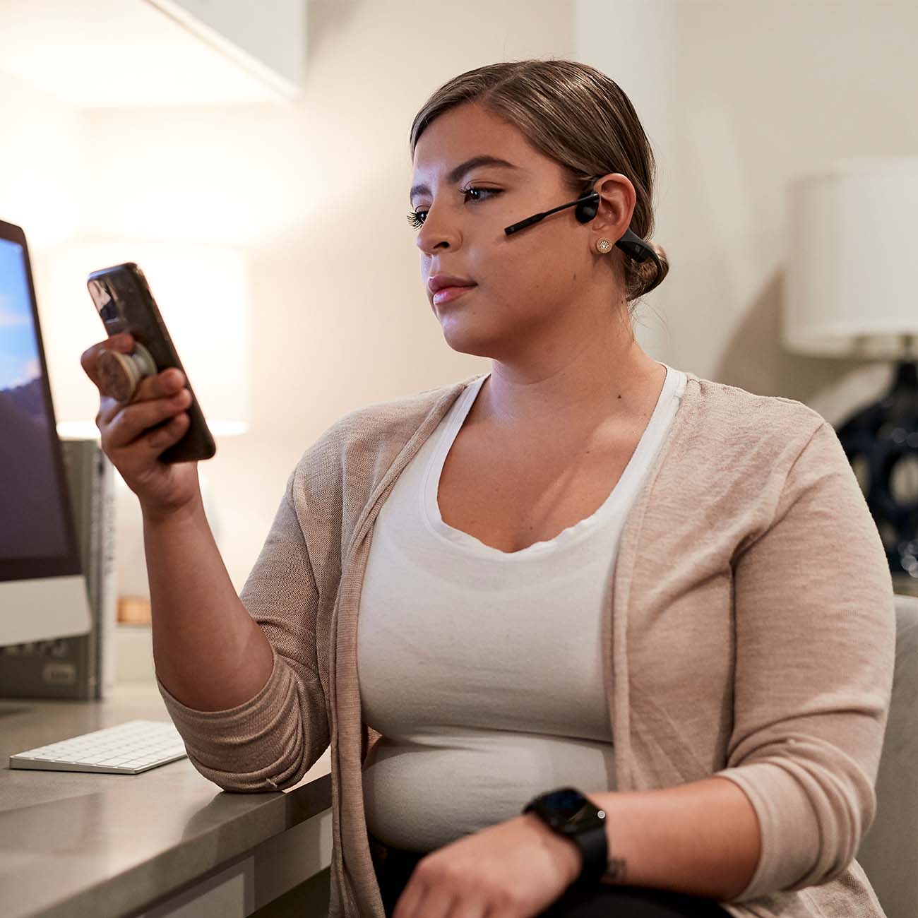 A person wearing Boeings Aftershokz OpenComm Headphones sits at a desk, smartphone in hand. In the background, a computer monitor, keyboard, and lamp are visible. The open-ear design helps them stay focused on their phone while being aware of their surroundings.