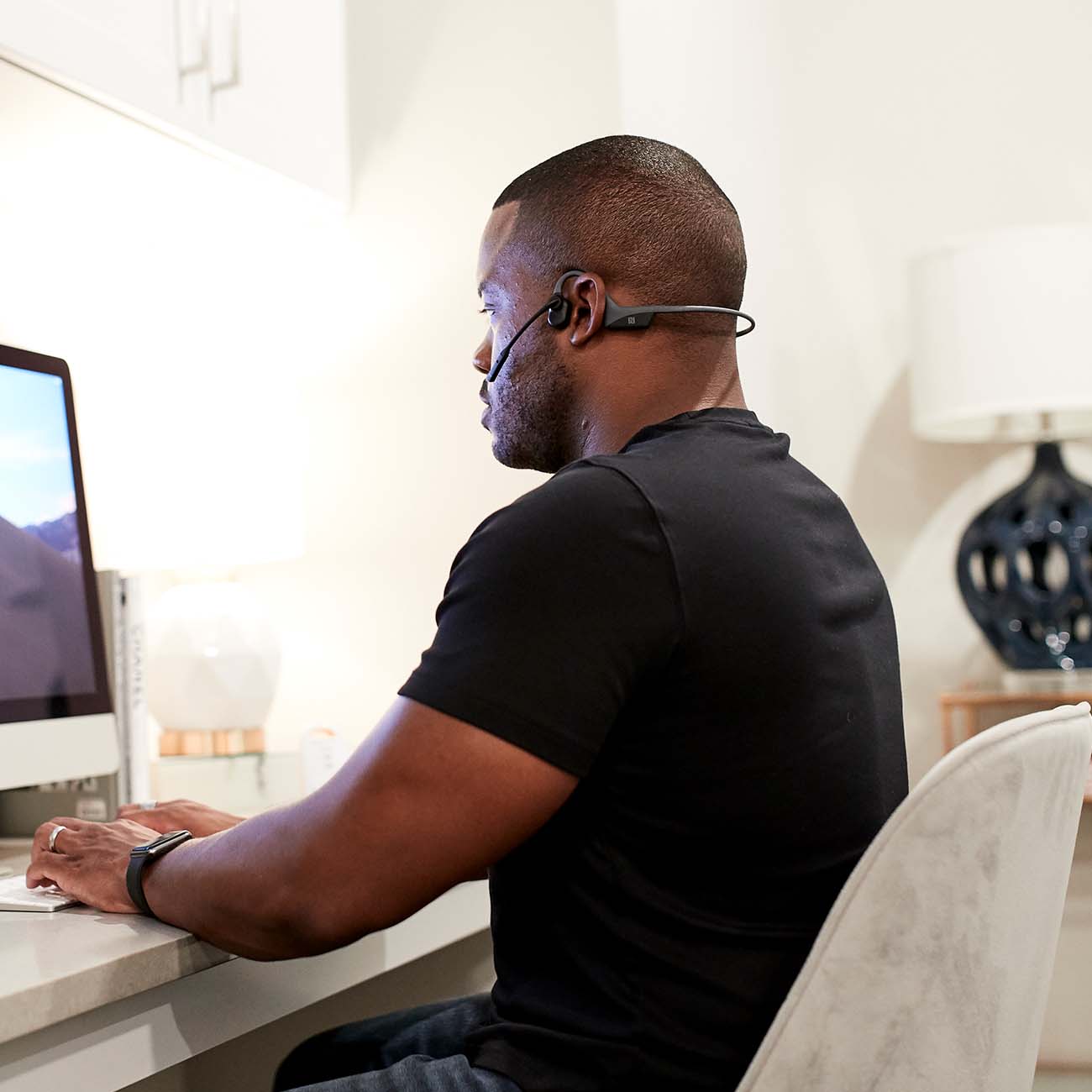 A man wearing a black T-shirt and Aftershokz OpenComm Headphones by Boeing sits at a desk, typing on a computer. A lamp and decorative item are visible in the background on the white desk.