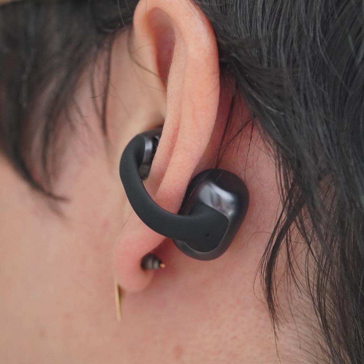 A close-up shot shows a person with short dark hair and a metal earring in their lobe wearing BOEING Shokz OpenDots Earbuds, which feature a sleek black, sweat-resistant design.