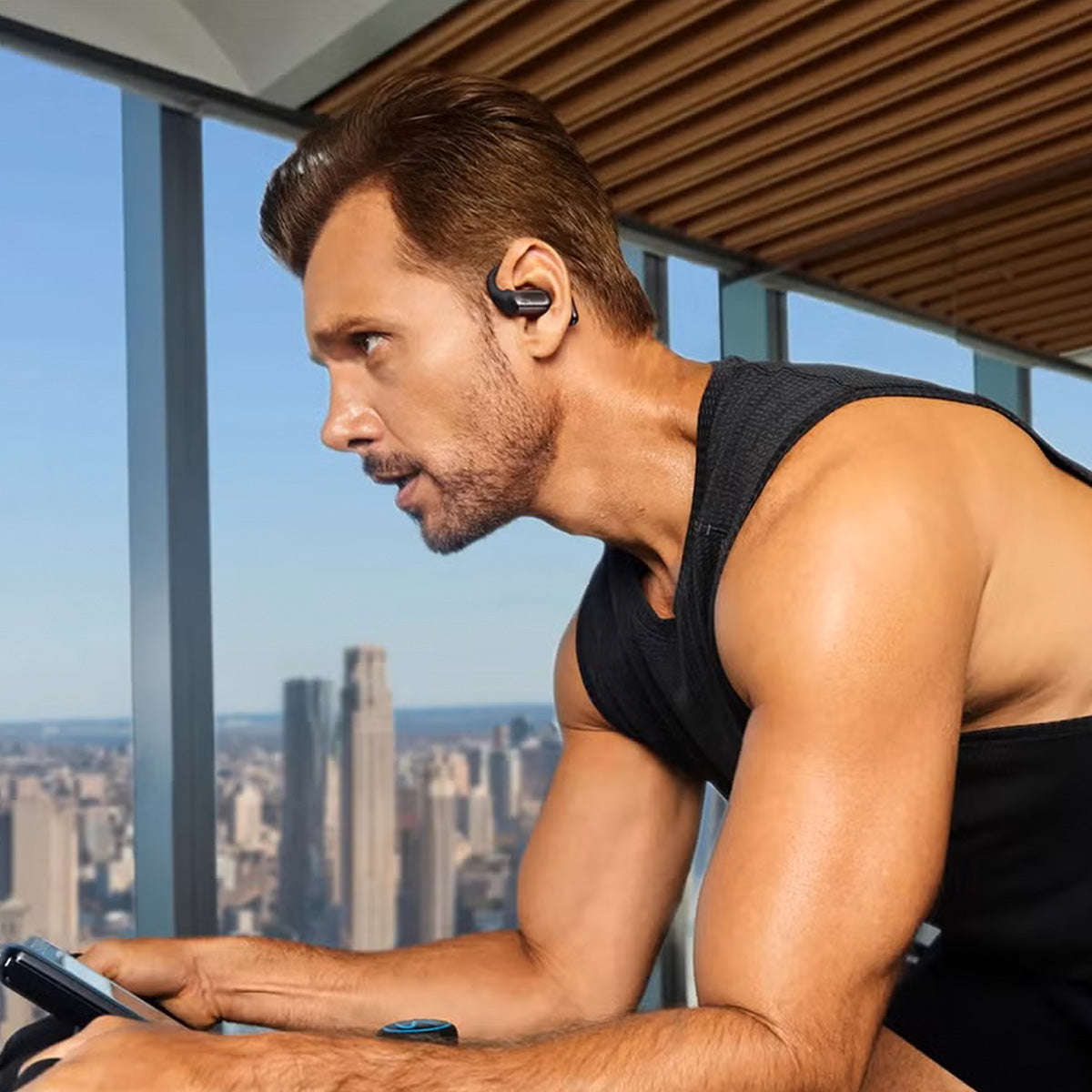Wearing BOEING Shokz OpenFit 2+ Earbuds and a sleeveless shirt, a man exercises on a stationary bike in a modern gym with large windows overlooking the city skyline, enjoying premium sound as he works out.