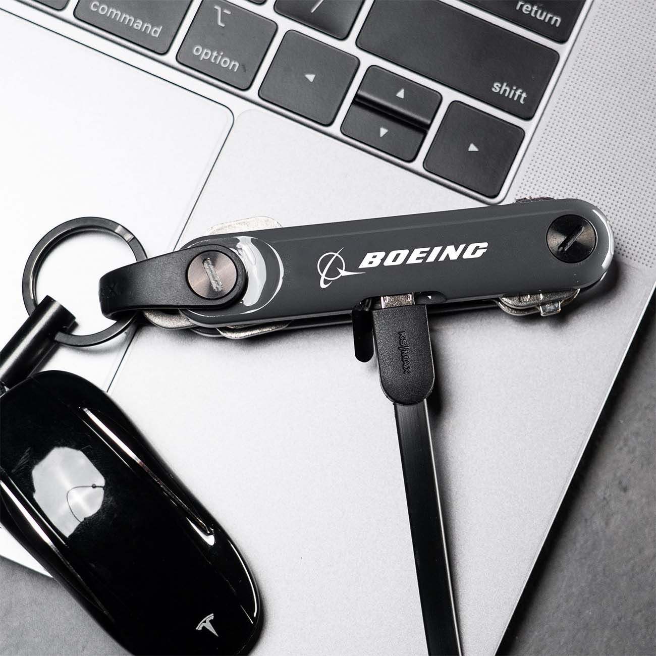 A Boeing Keysmart Max with a USB cable sits on a laptop keyboard, featuring Bluetooth capabilities. Nearby, a Tesla key fob is casually placed.
