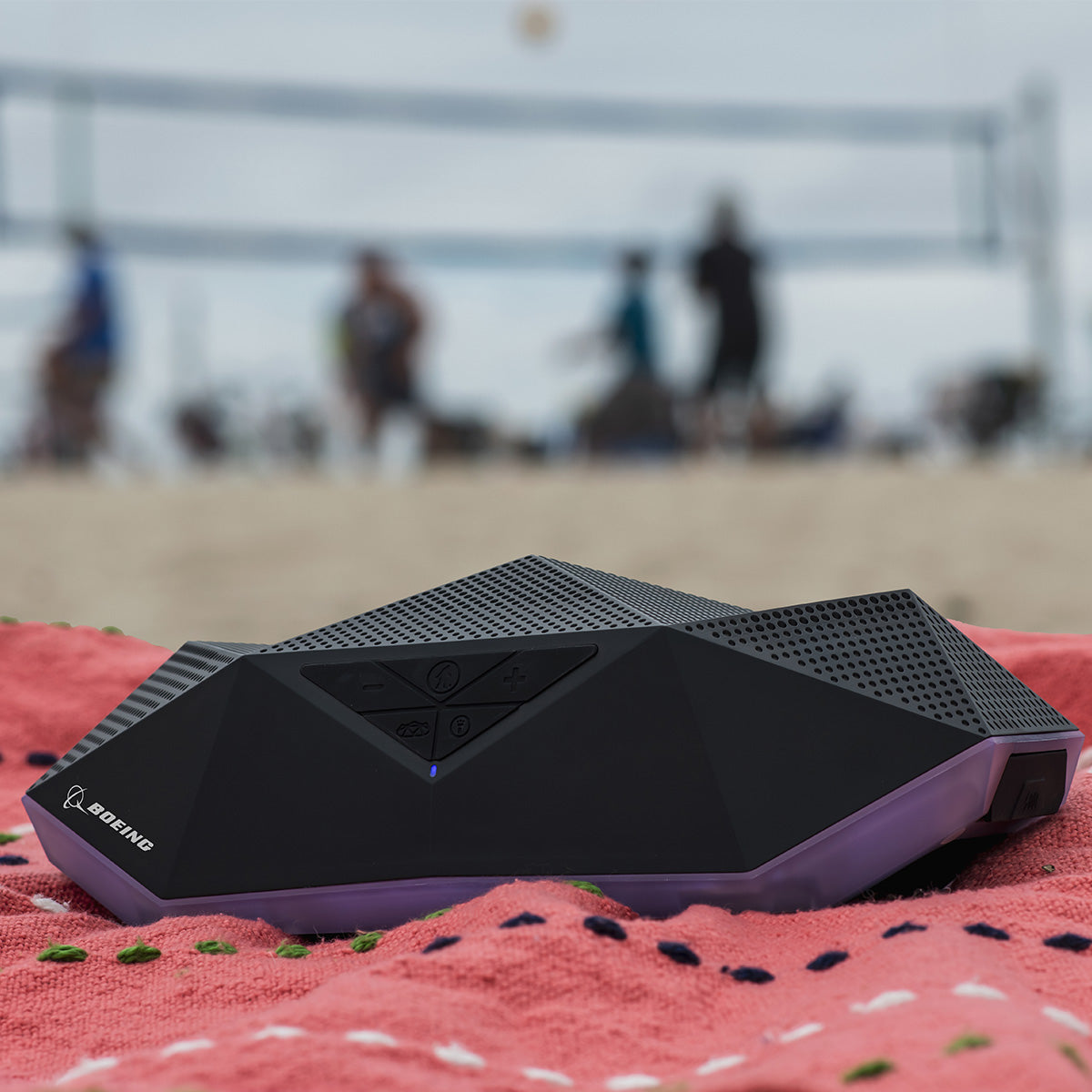 The black BOEING Outdoor Tech Boeing Rugged Range Speaker with 360° sound sits on a red blanket at the beach, while people play volleyball in the blurred background.