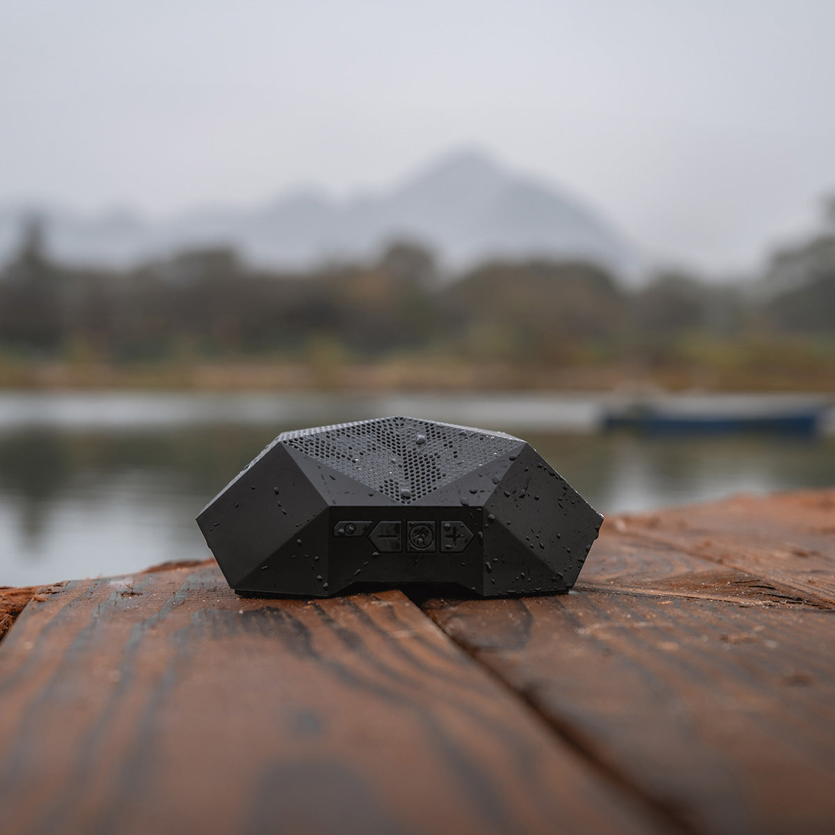 The BOEING Outdoor Tech Turtle Shell 4.0 Speaker, featuring a black geometric design and waterproof build, sits on a wet wooden surface by a lake with mountains and trees blurred in the cloudy background.