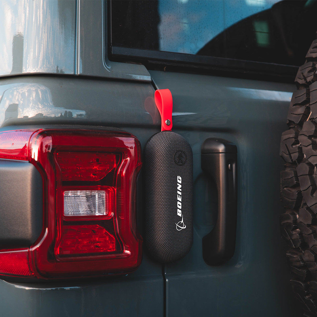 A black Outdoor Tech Boeing Bolt Wireless Speaker with a red strap and BOEING logo hangs from the rear door of a gray vehicle near the spare tire and taillight.