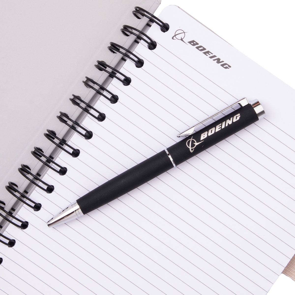 A black ballpoint pen from the Boeing V-22 Osprey Graphic Notebook Pen Set rests on an open spiral-bound notebook with lined pages.