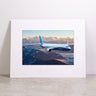 The Boeing 737-10 Matted Print - Large by Boeing features a high-resolution digital image of a blue Boeing airplane soaring over snow-covered mountains at sunset, beautifully framed and matted, and displayed on a gray surface with a plain background.