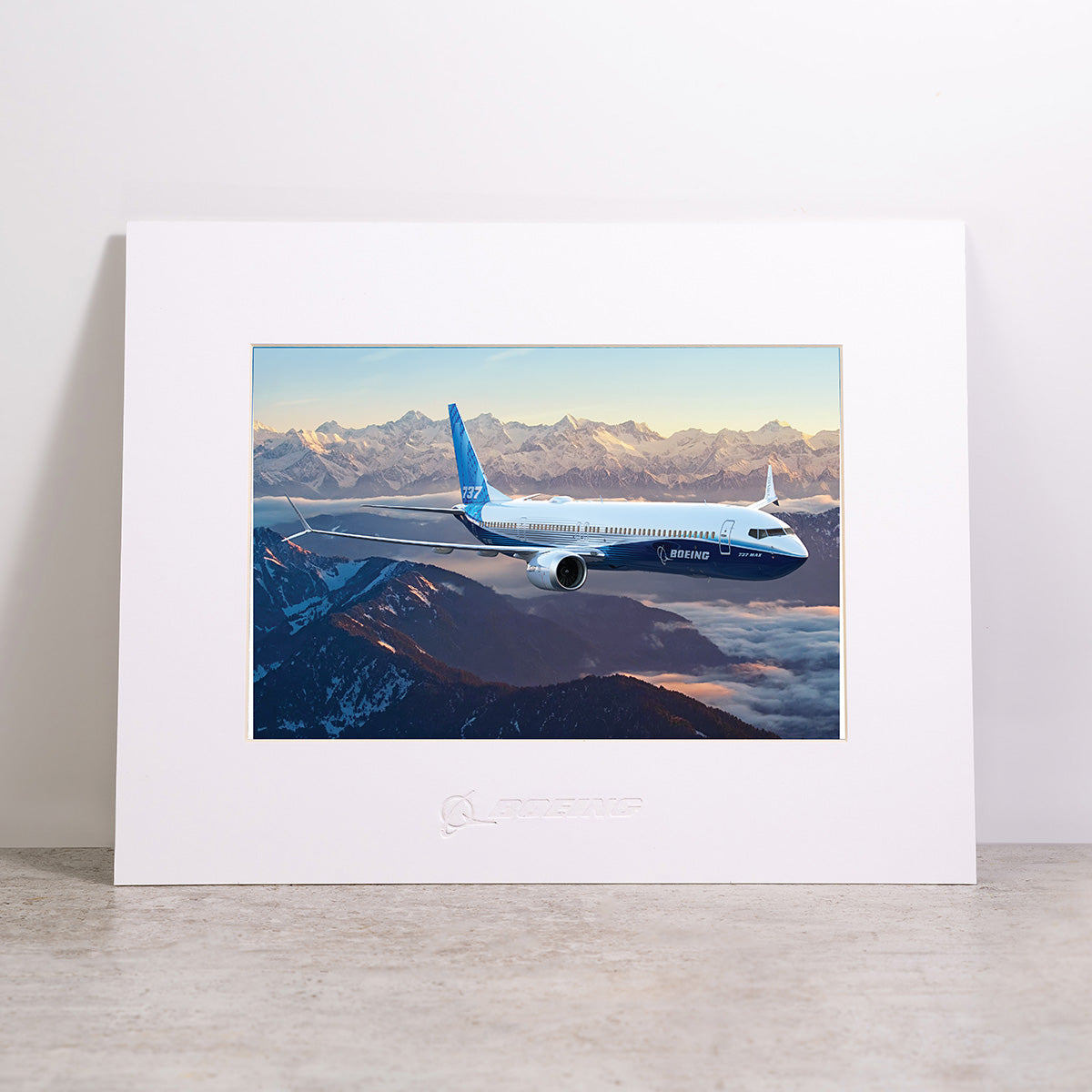 The Boeing 737-10 Matted Print - Large by Boeing features a high-resolution digital image of a blue Boeing airplane soaring over snow-covered mountains at sunset, beautifully framed and matted, and displayed on a gray surface with a plain background.