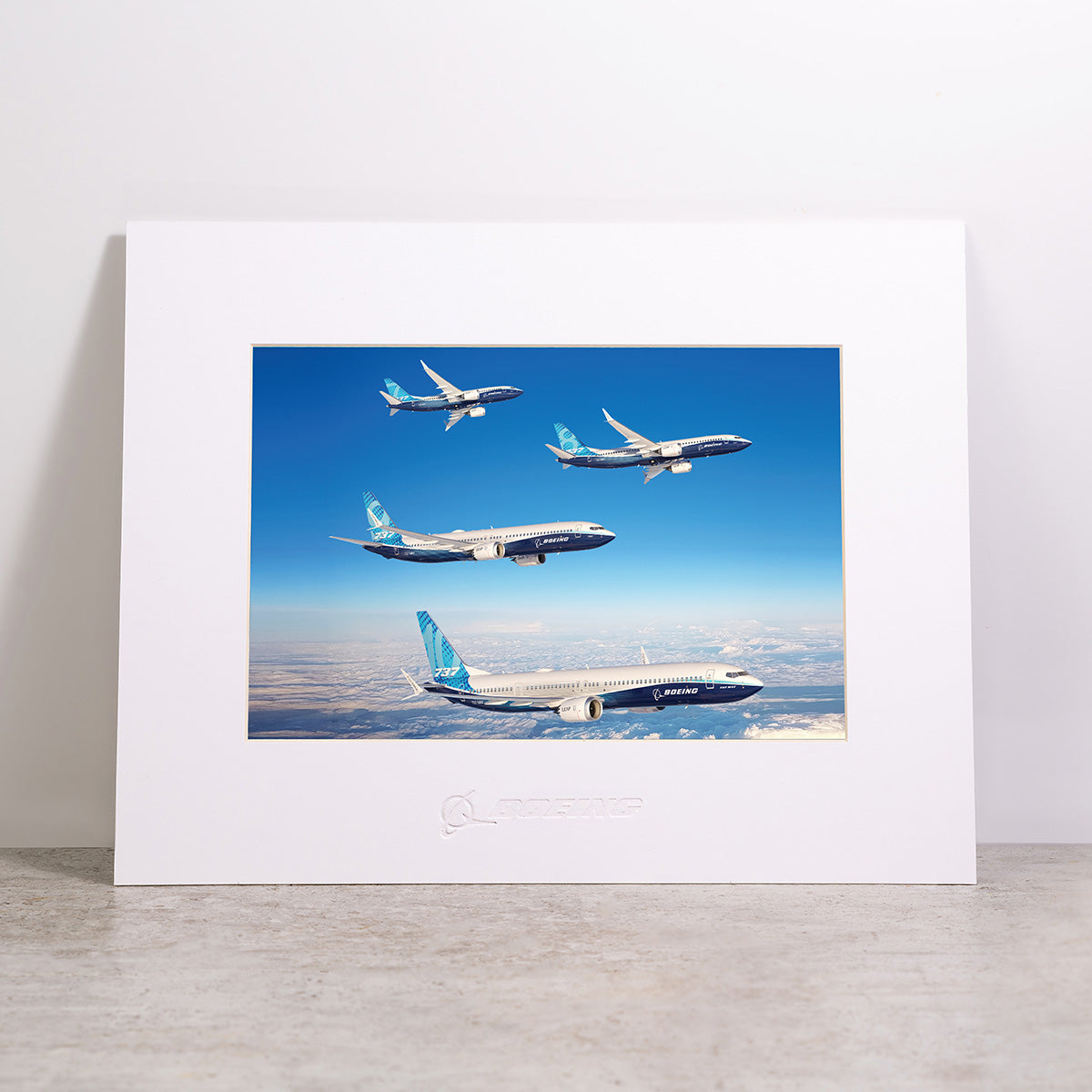 The Boeing 737 MAX Family Sky Matted Print - Small by Boeing is shown framed and matted, featuring four 737 MAX airplanes soaring against a blue sky with clouds. Displayed on a light-colored surface against a plain white wall, this high-resolution digital print delivers striking detail and clarity, and includes the iconic Boeing logo.