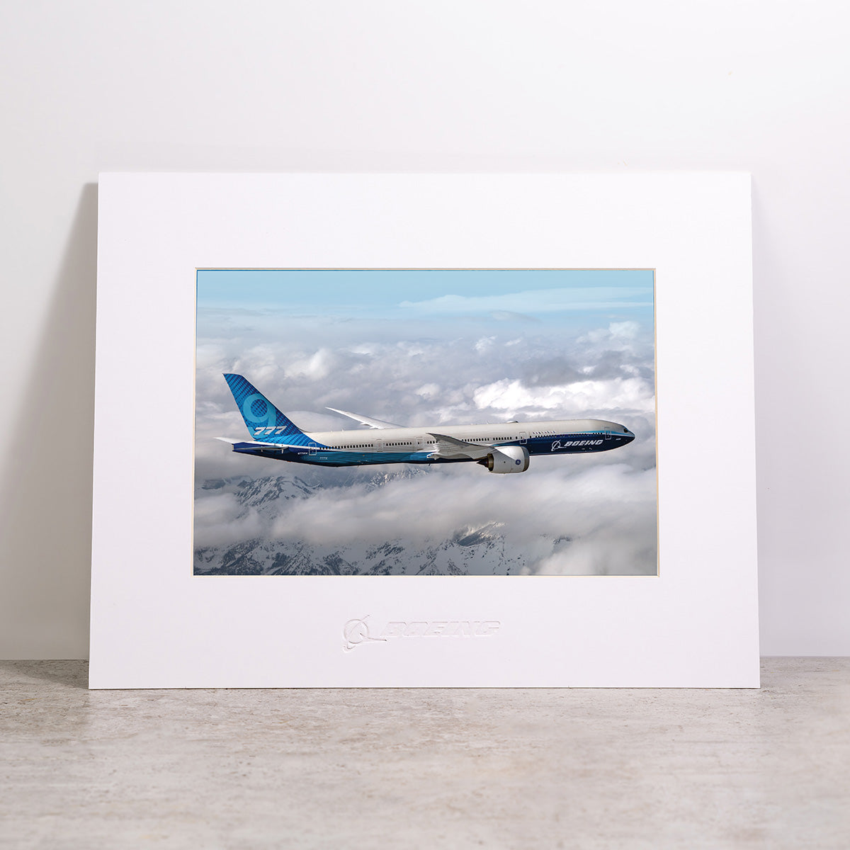 Introducing the Boeing 777-9 Matted Print - Large by Boeing: a framed, matted print featuring a vivid image of a blue Boeing 777-9 soaring above clouds and snowy mountains. Displayed upright on a gray surface against a plain light background, this high-resolution digital print showcases exceptional detail—making it an eye-catching addition for any aviation enthusiast.