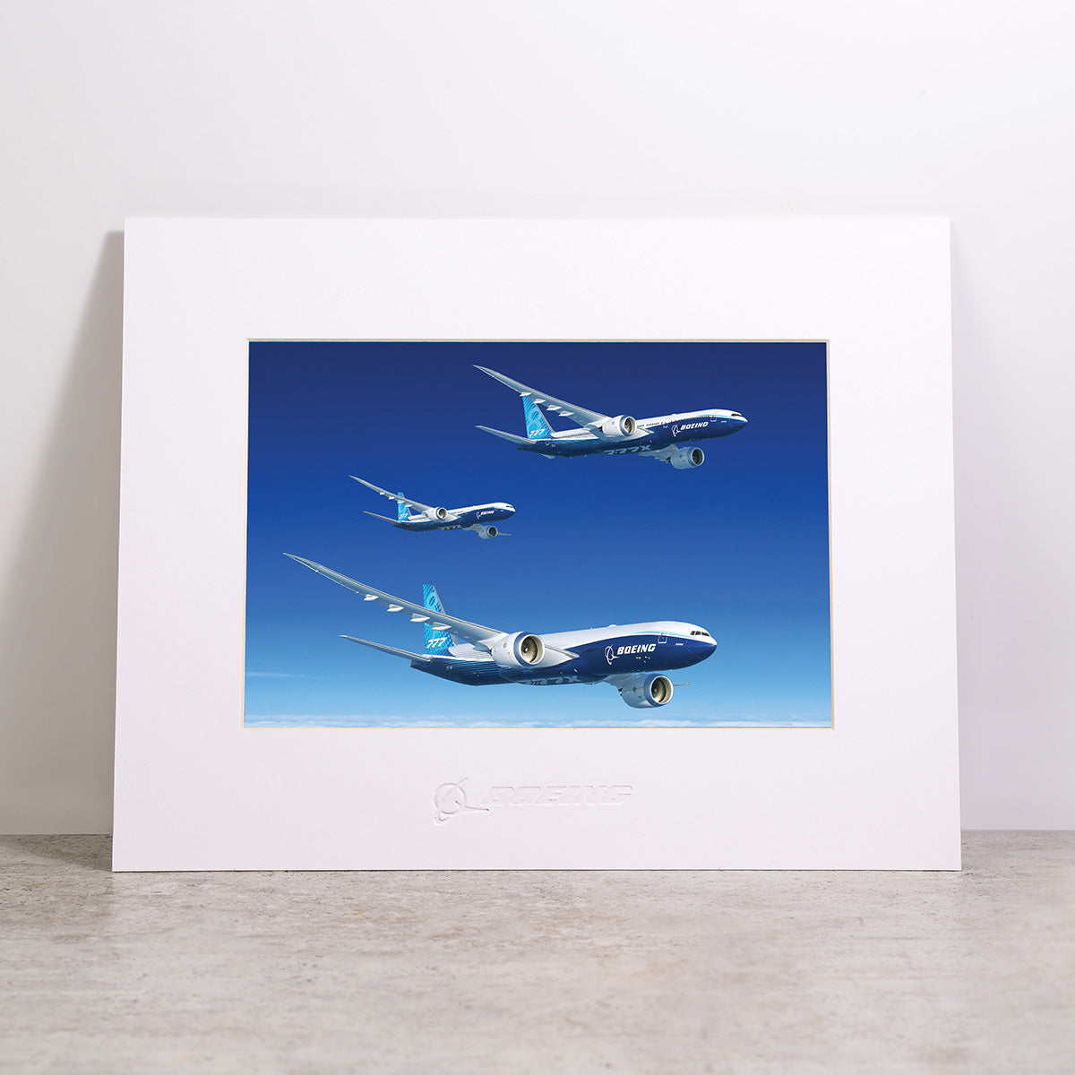 Framed and matted “Boeing 777X Family Matted Print - Large” by Boeing, featuring three blue Boeing airplanes soaring at varying heights against a clear blue background, with the Boeing logo embossed on the white mat.