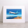 Here is the revised sentence using your product data: A Boeing 787-9 Matted Print - Small by Boeing, showcasing a high-resolution image of a Boeing 787 aircraft flying over a coastal landscape with blue sky, displayed upright on a light-colored surface against a plain white background.