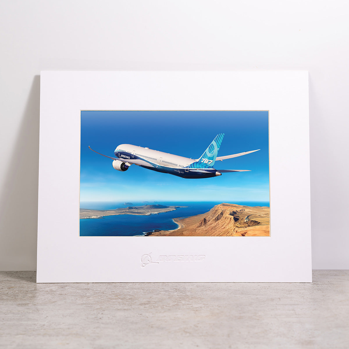 Here is the revised sentence using your product data: A Boeing 787-9 Matted Print - Small by Boeing, showcasing a high-resolution image of a Boeing 787 aircraft flying over a coastal landscape with blue sky, displayed upright on a light-colored surface against a plain white background.