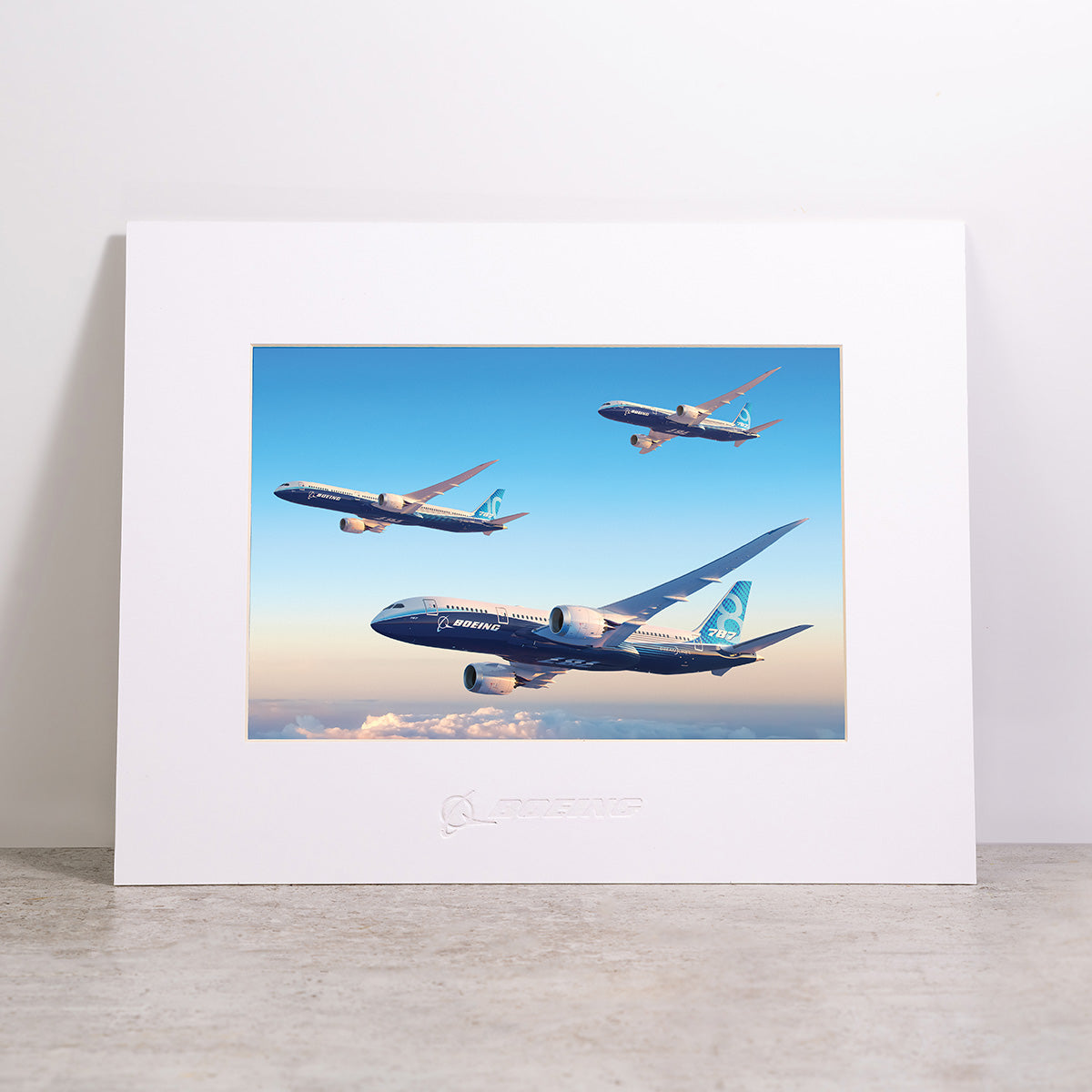 Showcase your love for aviation with the Boeing 787 Dreamliner Family Matted Print - Large by Boeing. This 16 x 20 wall art features a high-resolution digital image of three Boeing airplanes soaring above the clouds against a vivid blue sky. The matted print is elegantly presented in a frame, with the iconic Boeing logo embossed along the bottom, making it a striking addition to any aviation enthusiast’s collection.