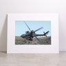 The Boeing AH-64 Matted Print - Small by Boeing is a framed, matted print featuring a military helicopter soaring above a rugged landscape. Displayed upright on a stone surface against a plain white background, this striking image is produced with high-resolution digital printing to ensure crisp detail and lasting quality.
