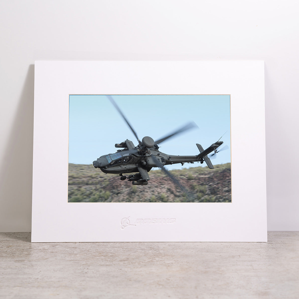 The Boeing AH-64 Matted Print - Large by Boeing features a high-resolution digital image of a military helicopter soaring above a landscape with blurred rotor blades, elegantly presented as a matted print in a frame and displayed on a gray surface against a light background.
