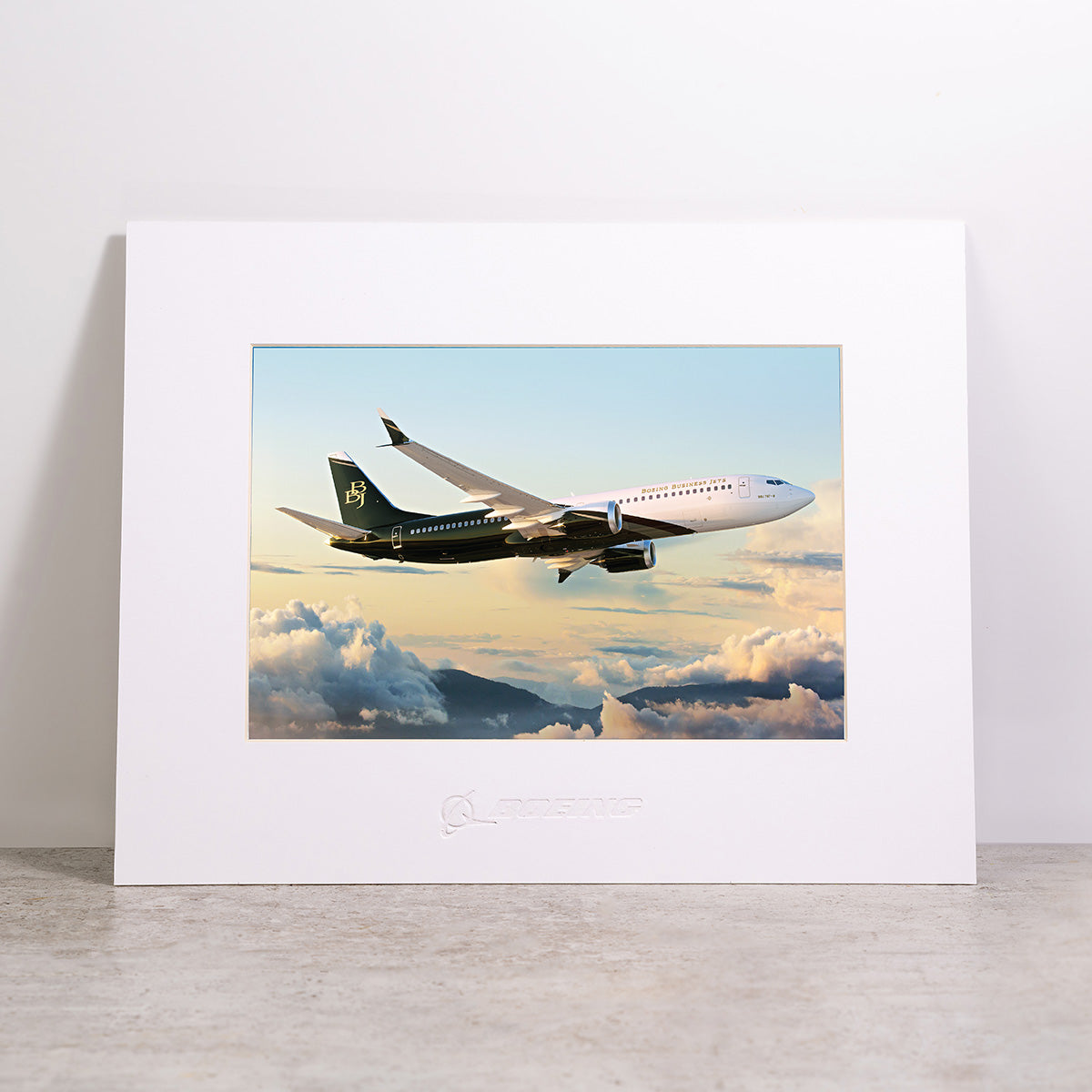 Boeing BBJ Matted Print - Small by Boeing, featuring a jet airplane soaring above clouds at sunset, showcases high-resolution digital printing for stunning detail and includes the Boeing logo embossed beneath the image.