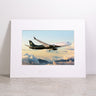 Showcasing the Boeing BBJ, the Boeing BBJ Matted Print - Large features a high-resolution image of a jet soaring above sunset clouds. Presented on a light-colored surface with a white mat embossed with the Boeing logo, this striking art print by Boeing offers vibrant detail and exceptional clarity through advanced digital printing.