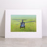 Displayed upright against a white wall and resting on a concrete surface, the Boeing CH-47F Block II Matted Print - Small by Boeing features a framed photo of this iconic helicopter flying low over a grassy field. This striking matted print showcases high-resolution digital printing, ensuring every detail is crisp and vivid.