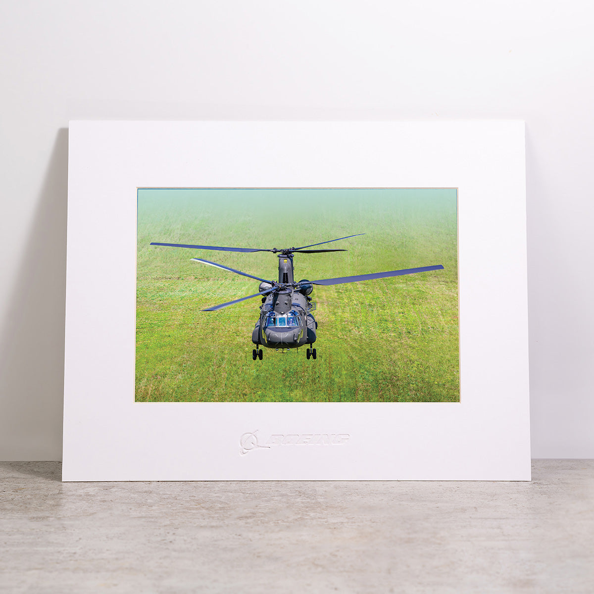 Displayed upright against a white wall and resting on a concrete surface, the Boeing CH-47F Block II Matted Print - Small by Boeing features a framed photo of this iconic helicopter flying low over a grassy field. This striking matted print showcases high-resolution digital printing, ensuring every detail is crisp and vivid.