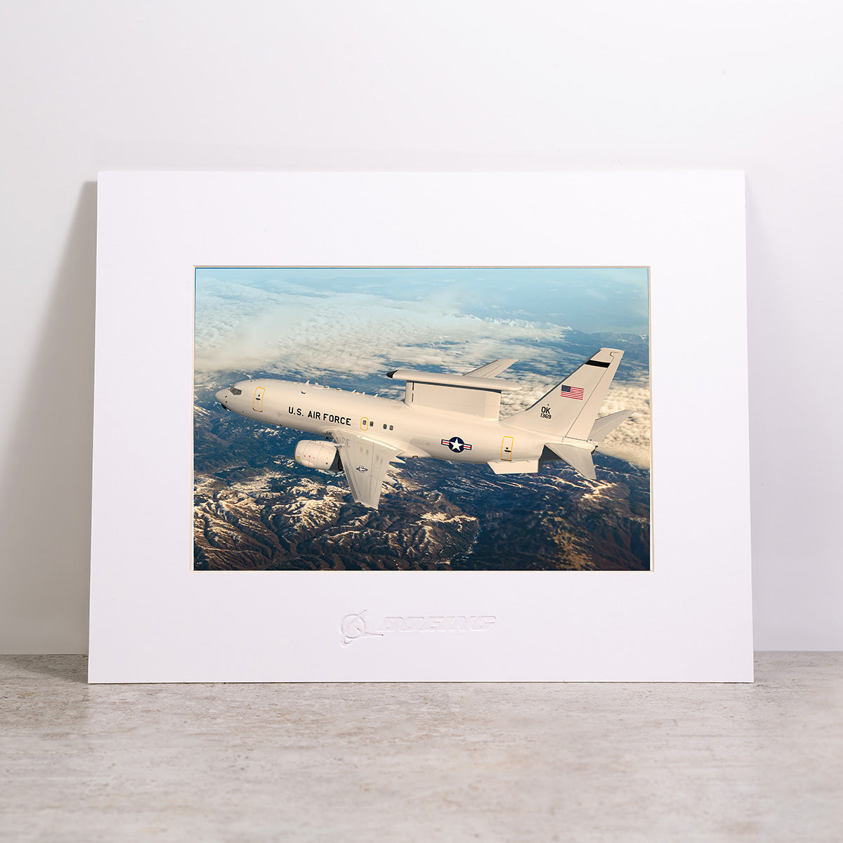The Boeing E-7 AEW&C Matted Print – Large features a striking image of a U.S. Air Force aircraft soaring above clouds and land, with the Boeing logo embossed on the white mat for an elegant finish.