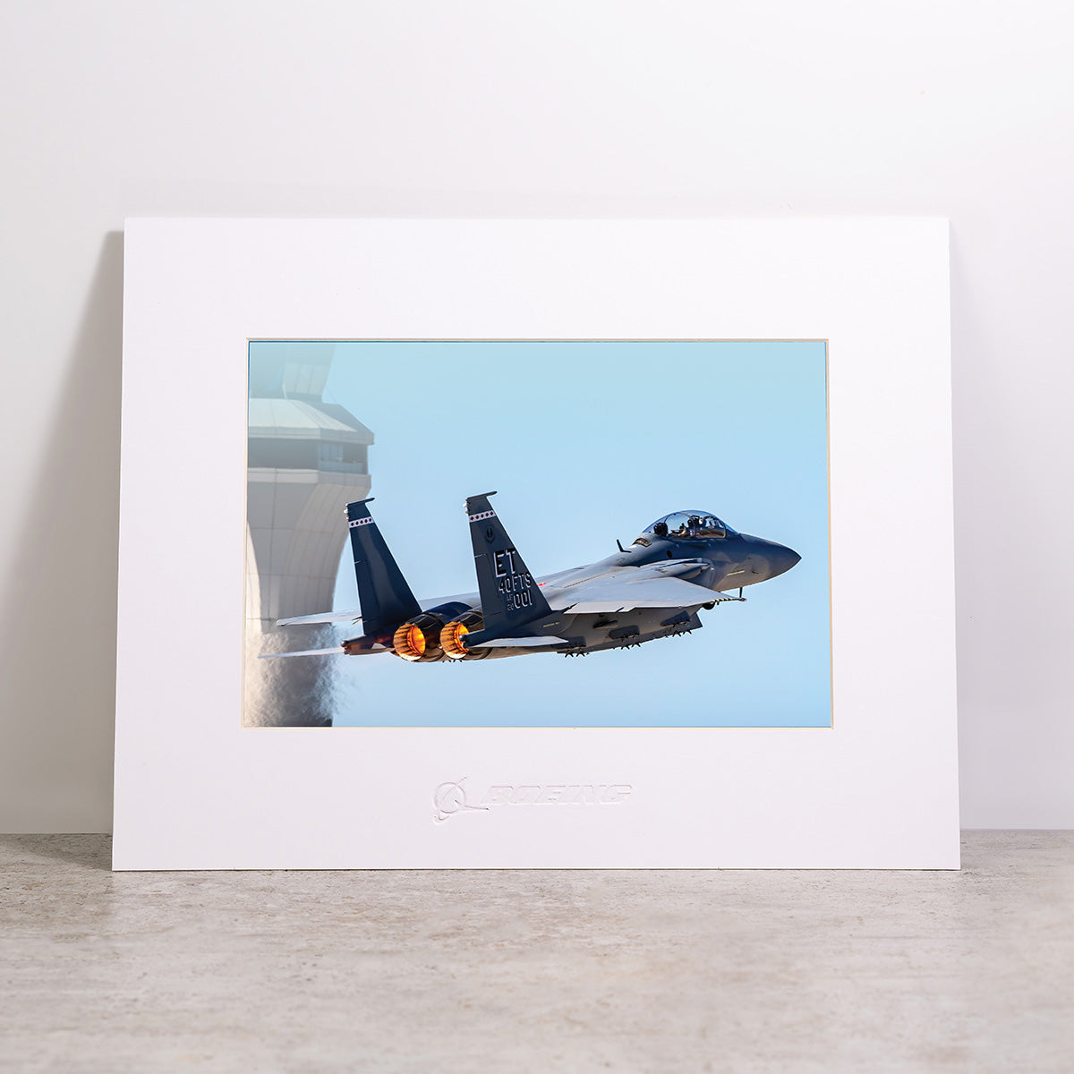 Display the Boeing F-15EX Eagle II Matted Print - Small by Boeing upright on a light-colored surface. This striking image features a fighter jet taking off with afterburners lit as it soars past an air traffic control tower against a clear blue sky. The high-resolution digital printing ensures crisp detail and vibrant color throughout the matted print.