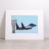 A Boeing F-15EX Eagle II Matted Print - Large by Boeing, featuring a high-resolution digital image of the fighter jet taking off with afterburners blazing against a blue sky and control tower backdrop, presented in a matted frame and displayed on a gray surface.