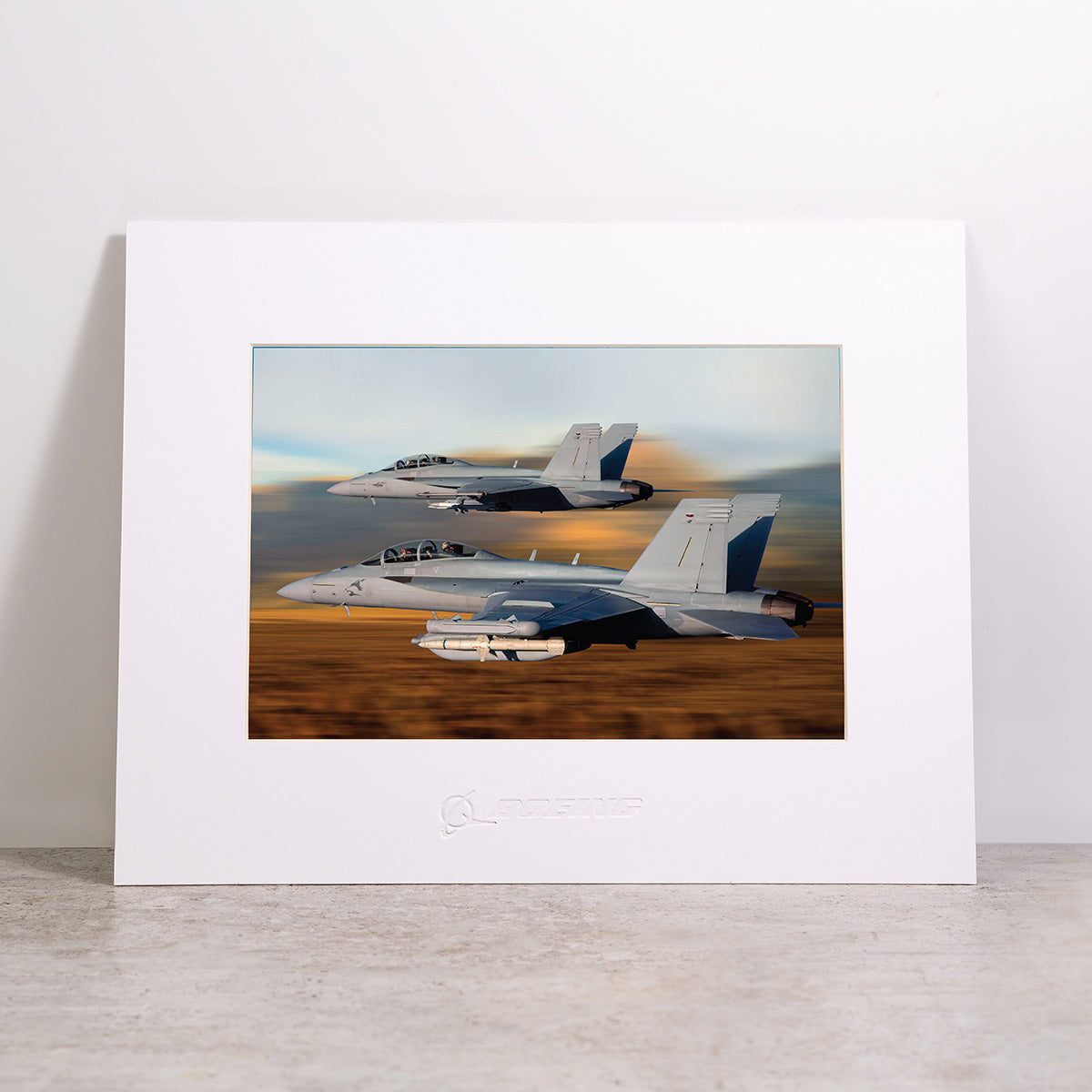 The Boeing F/A-18 & EA-18G Matted Print - Small by Boeing features a high-resolution framed digital print of two military fighter jets soaring in formation over a blurred landscape, surrounded by a white mat with the Boeing logo centered below the image.