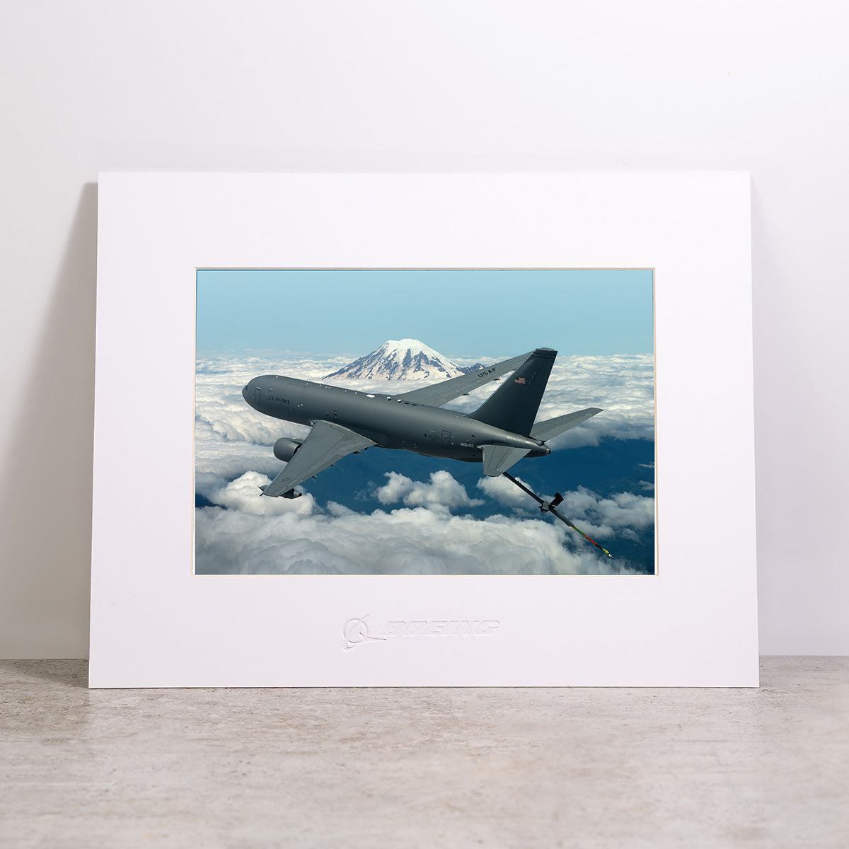 The Boeing KC-46A Pegasus Matted Print - Large by Boeing features a high-resolution digital image of a gray military aircraft soaring above clouds with a snow-capped mountain in the background, presented as a framed, matted print and displayed against a light-colored wall.