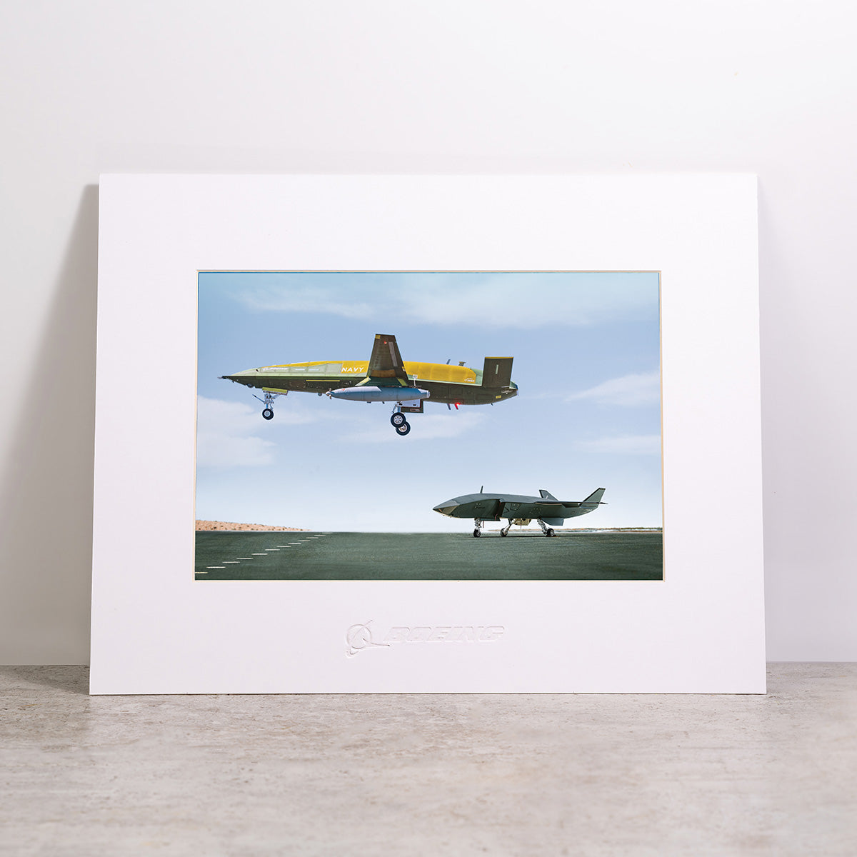 Showcased in a white frame on a gray surface, the Boeing MQ-25A & MQ-28 Matted Print - Large features vivid, high-resolution digital imagery of one unmanned aircraft in flight above a runway while another remains on the ground beneath a clear blue sky. Expertly produced by Boeing, this print combines meticulous craftsmanship with striking aviation photography.