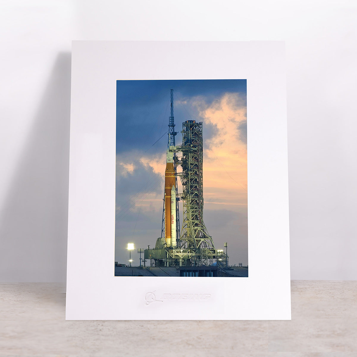 The Boeing SLS Matted Print - Small by Boeing features a high-resolution framed photo of a rocket on the launch pad at sunset, with dramatic clouds in the sky. Displayed upright on a light-colored surface against a plain background, this stunning digital print offers crisp detail, making it an ideal choice for display as an impressive matted print.