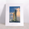 Introducing the Boeing SLS Matted Print - Large by Boeing: a framed, matted print featuring the towering SLS rocket on its launchpad at sunset beneath a dramatic sky. Expertly crafted with high-quality digital printing, this artwork arrives in a sleek frame displayed on a light-colored surface against a plain wall.