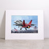 The Boeing T-7A Matted Print - Large by Boeing features a vivid, high-quality digital image of a red and gray jet soaring over an American flag, its afterburner blazing against a blue sky. Presented on a stone surface and elegantly framed with a white matte, this striking print showcases exceptional color and detail.