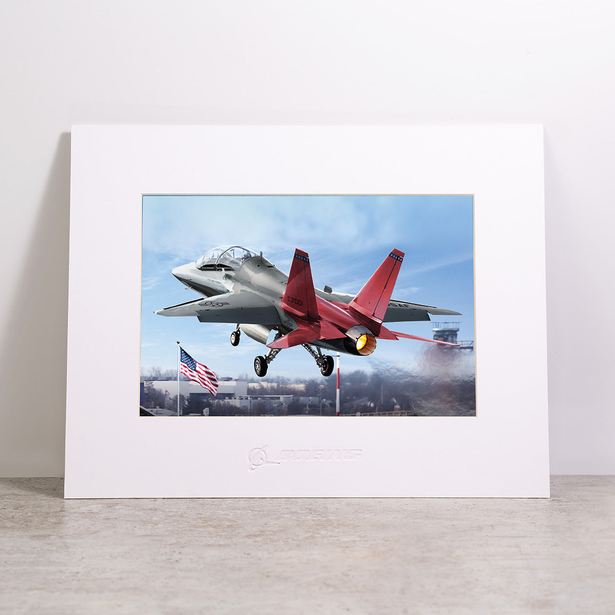 The Boeing T-7A Matted Print - Large by Boeing features a vivid, high-quality digital image of a red and gray jet soaring over an American flag, its afterburner blazing against a blue sky. Presented on a stone surface and elegantly framed with a white matte, this striking print showcases exceptional color and detail.