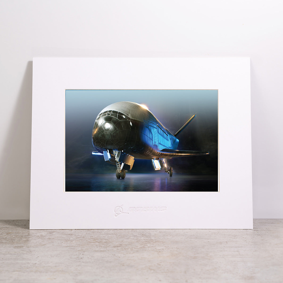 The Boeing X-37B Matted Print – Small by Boeing features a high-resolution digital image of the futuristic X-37B aircraft with its distinctive rounded nose, blue-lit accents, and extended landing gear. Expertly produced to capture every detail, this premium matted print is beautifully presented and displayed on a light surface against a plain white wall.