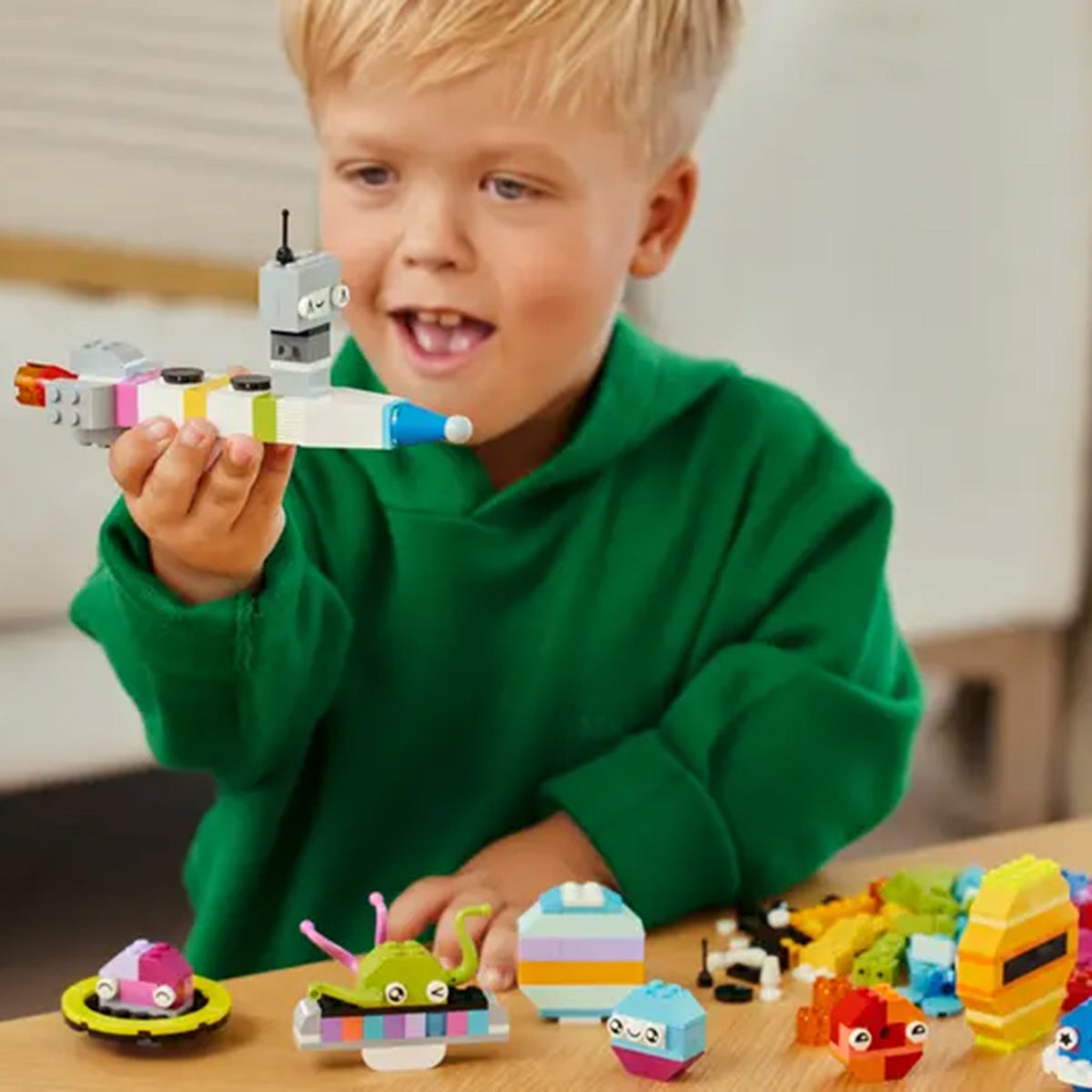 A young child in a green sweatshirt smiles while playing indoors with LEGO® Creative Space Planets, building colorful animals, vehicles, and a space shuttle featuring glow-in-the-dark bricks.