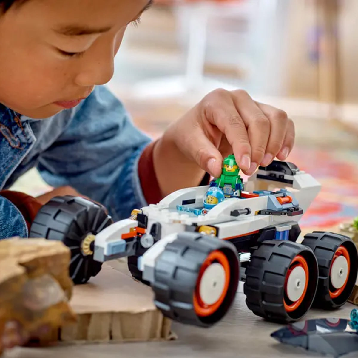 A child plays with the LEGO® Space Explorer Rover by LEGO, placing an astronaut mini-figure into the six-wheeled vehicle on a wooden surface with rocks nearby.