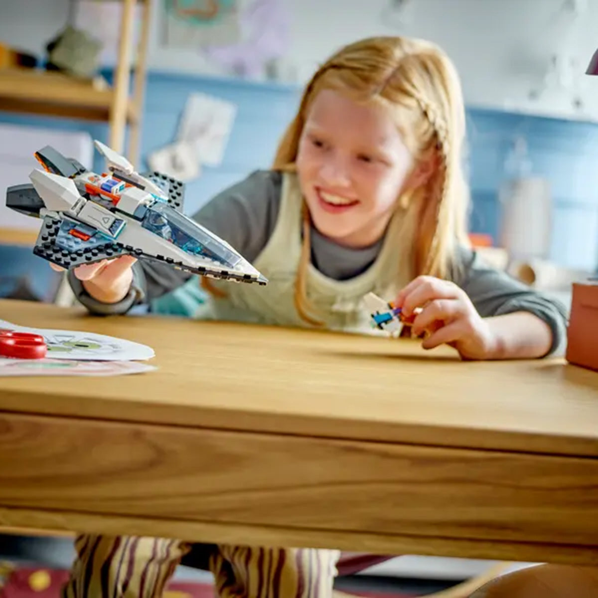 A smiling young girl with red hair plays at a wooden table, holding the LEGO® Interstellar Spaceship by LEGO, imagining space adventures in a cozy, softly lit room.