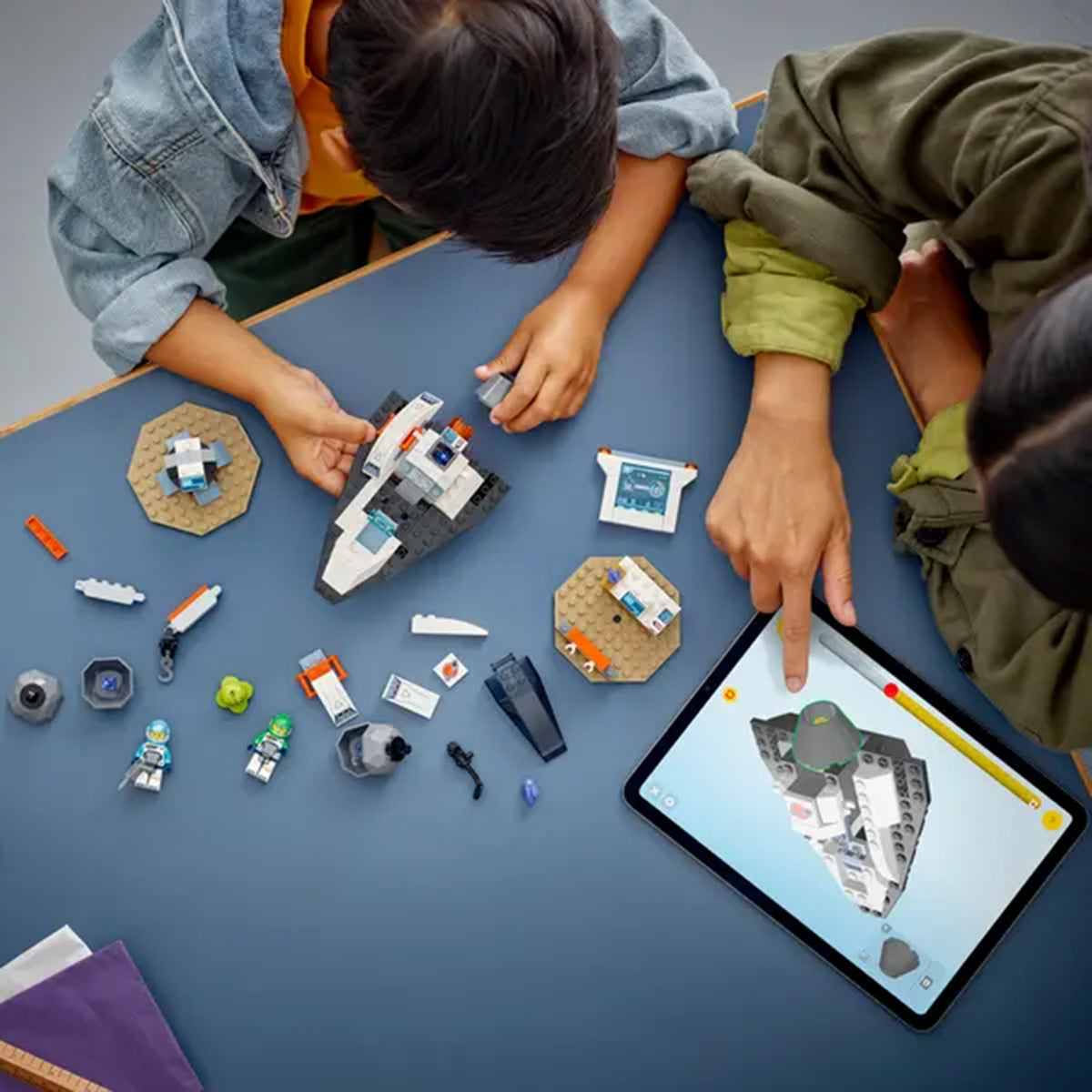 Two children build the LEGO® Spaceship and Asteroid Set by LEGO, following digital instructions on a tablet. Pieces from the set are spread around them as they work together on their cosmic project.