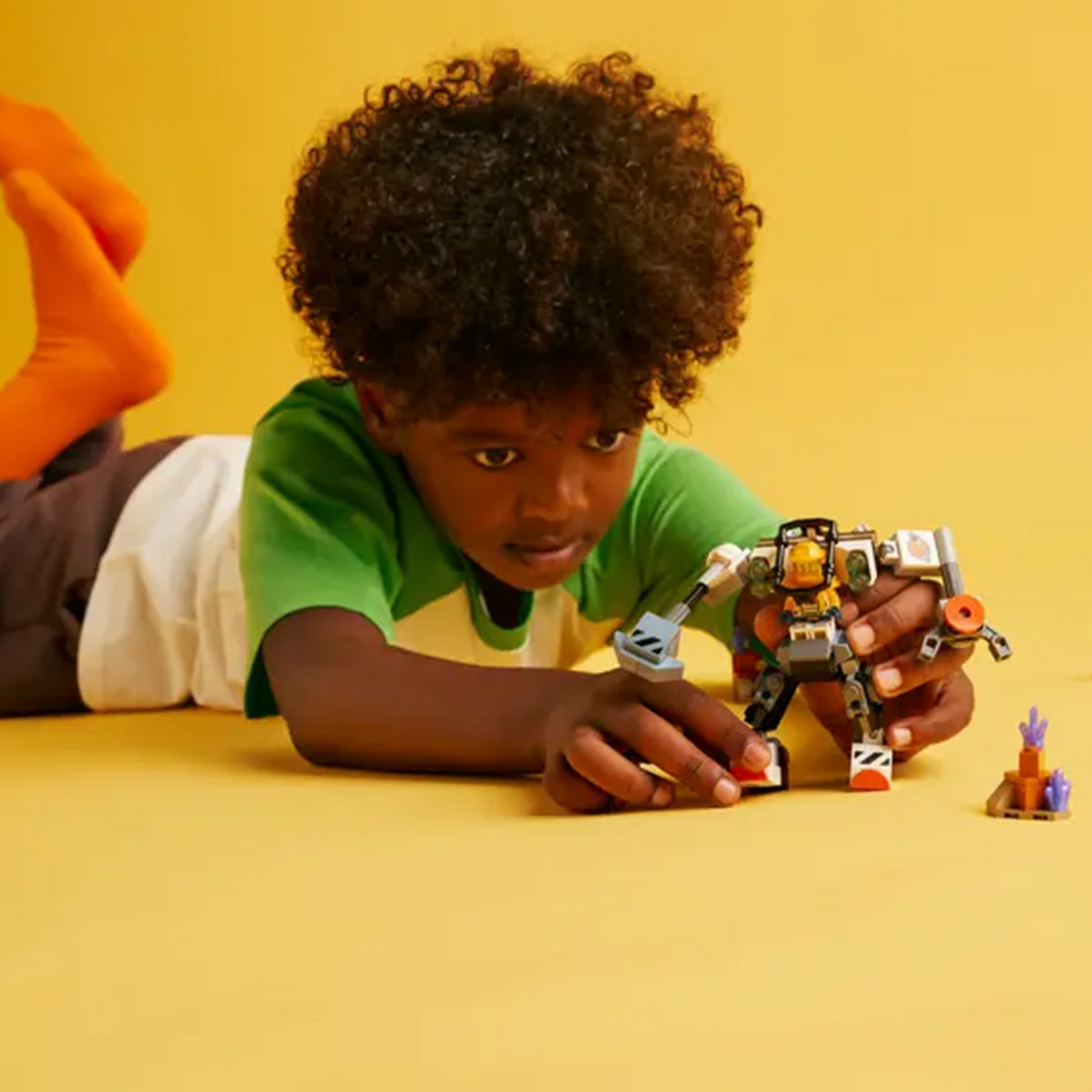 A young child with curly hair lies on a yellow floor, wearing an orange sock and green-and-white shirt, playing with the LEGO® Space Construction Mech by LEGO and small figures, ready for space adventures against a bright yellow background.