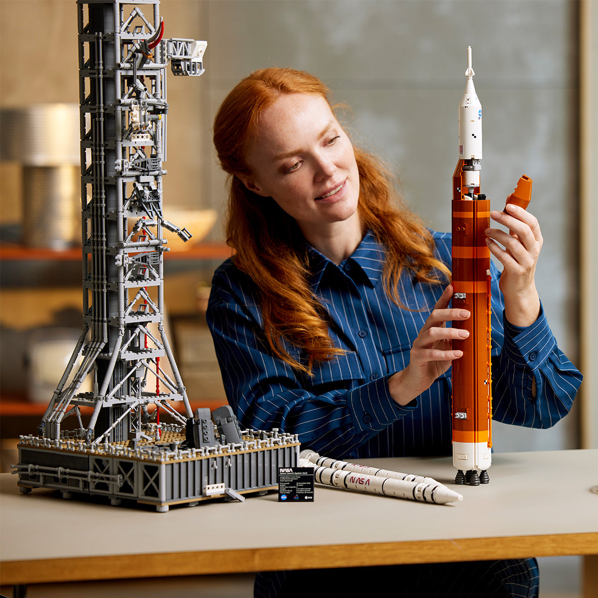 A woman with long red hair smiles as she builds the LEGO® NASA Artemis SLS Set by LEGO at a table, attaching a piece to the detailed orange and white multistage rocket and launch tower.