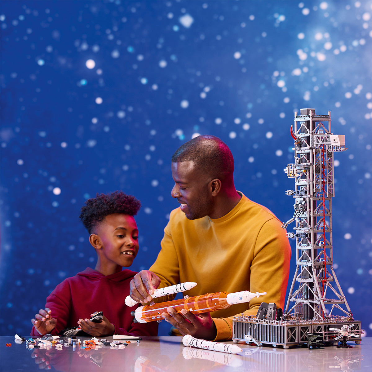 A man and a child smile as they assemble the LEGO® NASA Artemis SLS Set by LEGO at a table, with a starry, space-themed blue background behind them. Both are engaged in building the detailed multistage rocket together.