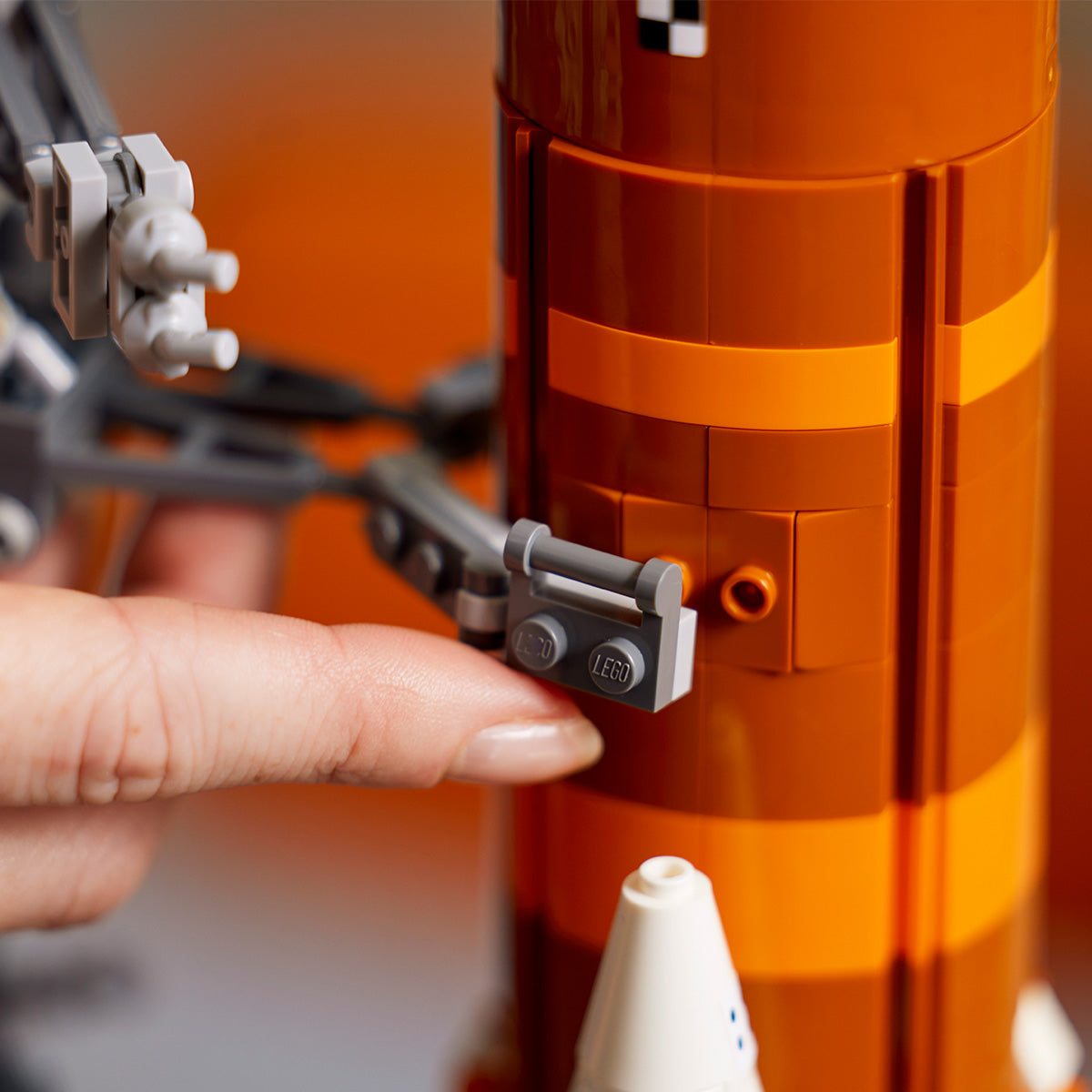 A close-up of a hand attaching a grey LEGO piece to an orange and brown LEGO® NASA Artemis SLS Set rocket model, featuring detailed, textured bricks inspired by the Artemis multistage launch system.