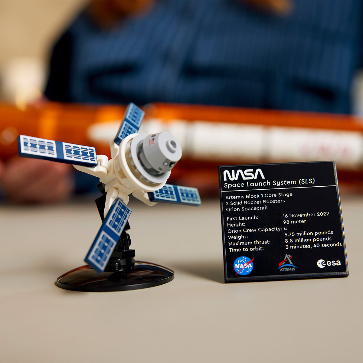 A LEGO® NASA Artemis SLS Set by LEGO stands on a table with a plaque showing Artemis I launch details, while blurred hands in the background capture the excitement surrounding this impressive multistage rocket model.