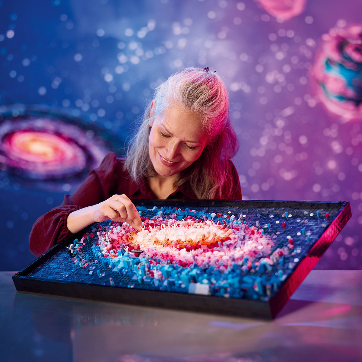 A smiling woman arranges mosaic pieces on a tray, creating a galaxy design inspired by the LEGO® The Milky Way Galaxy set by LEGO, with a vibrant, space-themed background filled with swirling galaxies.
