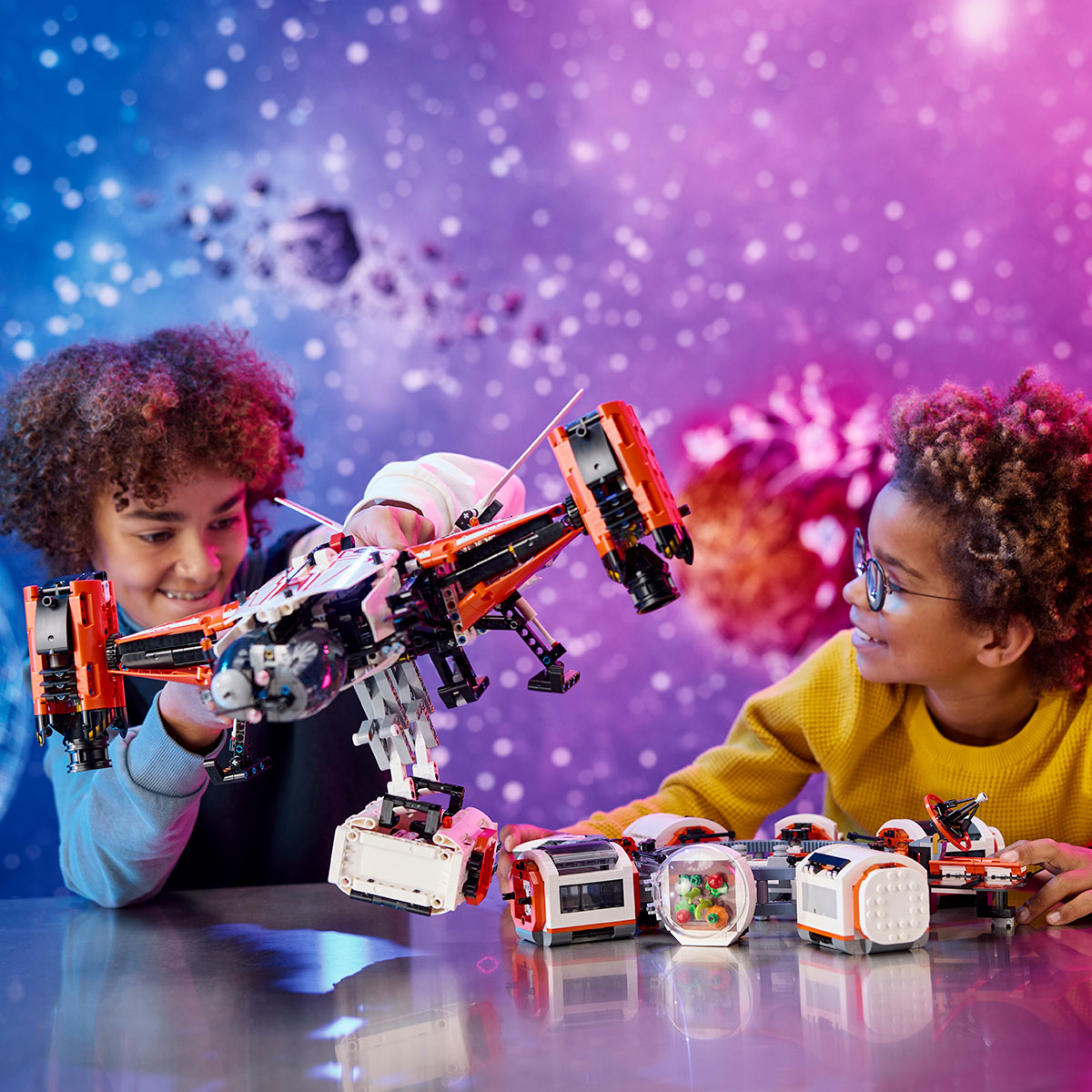 Two kids play with the LEGO® VTOL Heavy Cargo Spaceship LT81 and other space-themed LEGO Technic sets on a shiny table, as a colorful backdrop of stars and asteroids creates an adventurous outer space scene.