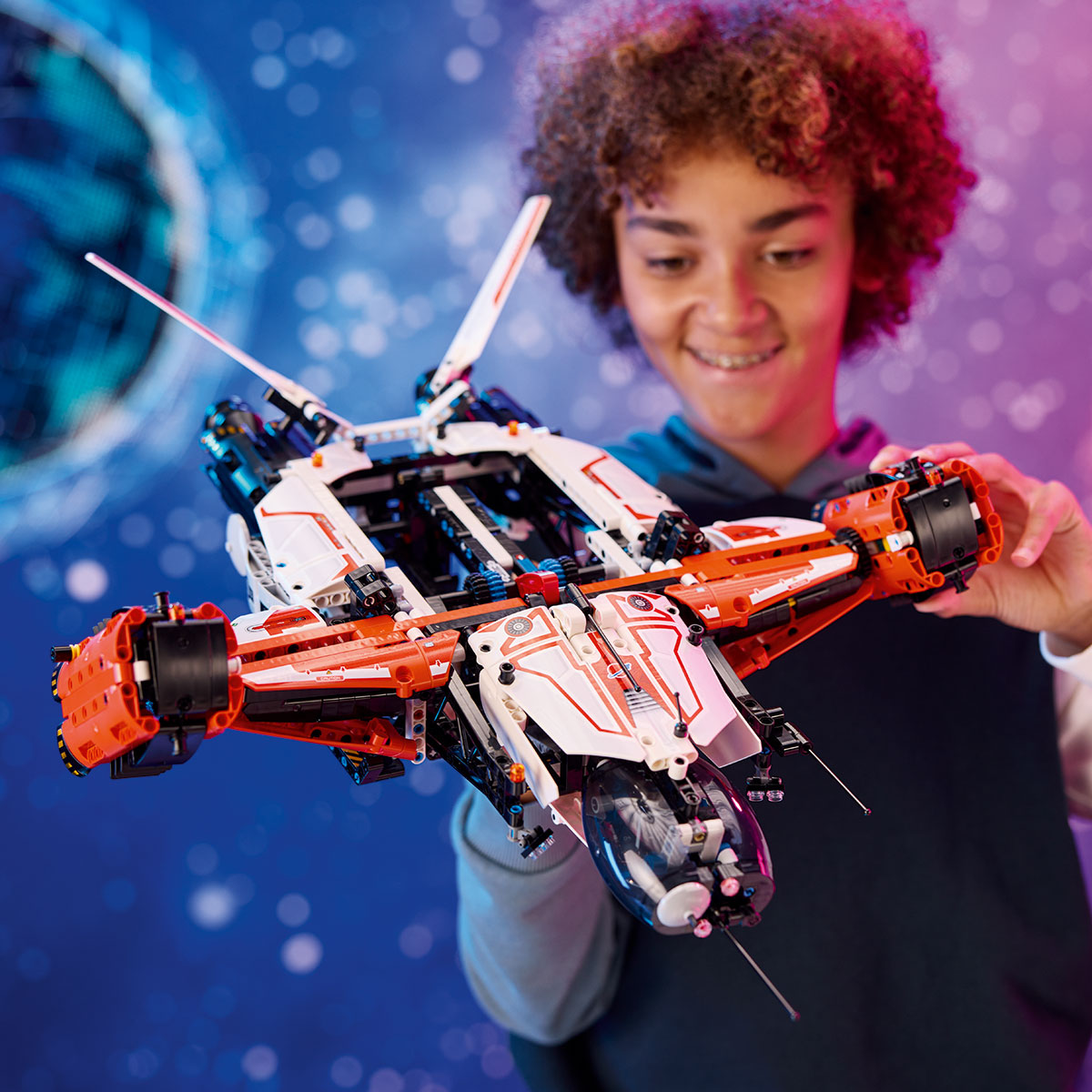 A smiling child holds the LEGO® VTOL Heavy Cargo Spaceship LT81 by LEGO, featuring orange, white, and black details, in front of a blue space-themed backdrop. A small space rover sits close by.